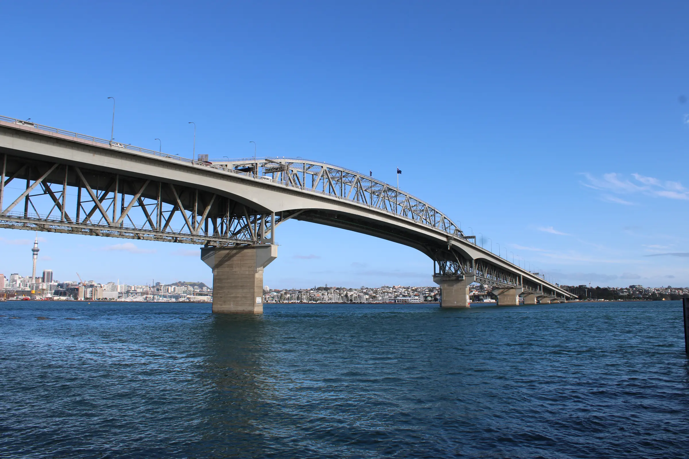 The Auckland Harbour Bridge from the water's edge on a clear blue day: the steel arch over the main span, its concrete piers in the Waitematā
