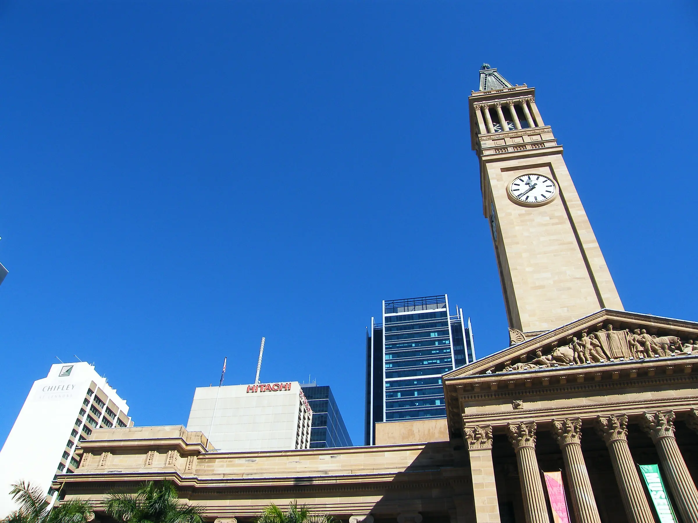 Brisbane City Hall's sandstone clock tower and columned portico over King George Square, office towers beside it, under a clear blue sky