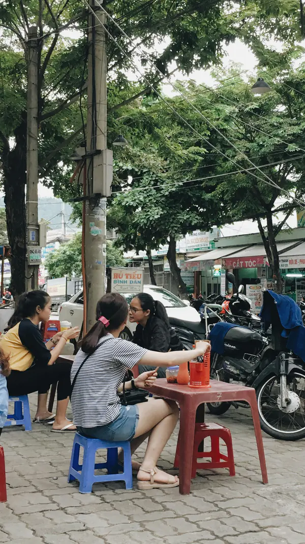 Friends on low plastic stools at a Đà Nẵng pavement cafe, drinks on a red table
