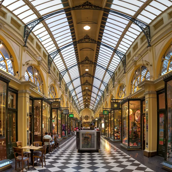 Inside a Melbourne arcade: a long Victorian shopping arcade under an arched glass roof, its shopfronts lit gold and its floor laid in black and white squares